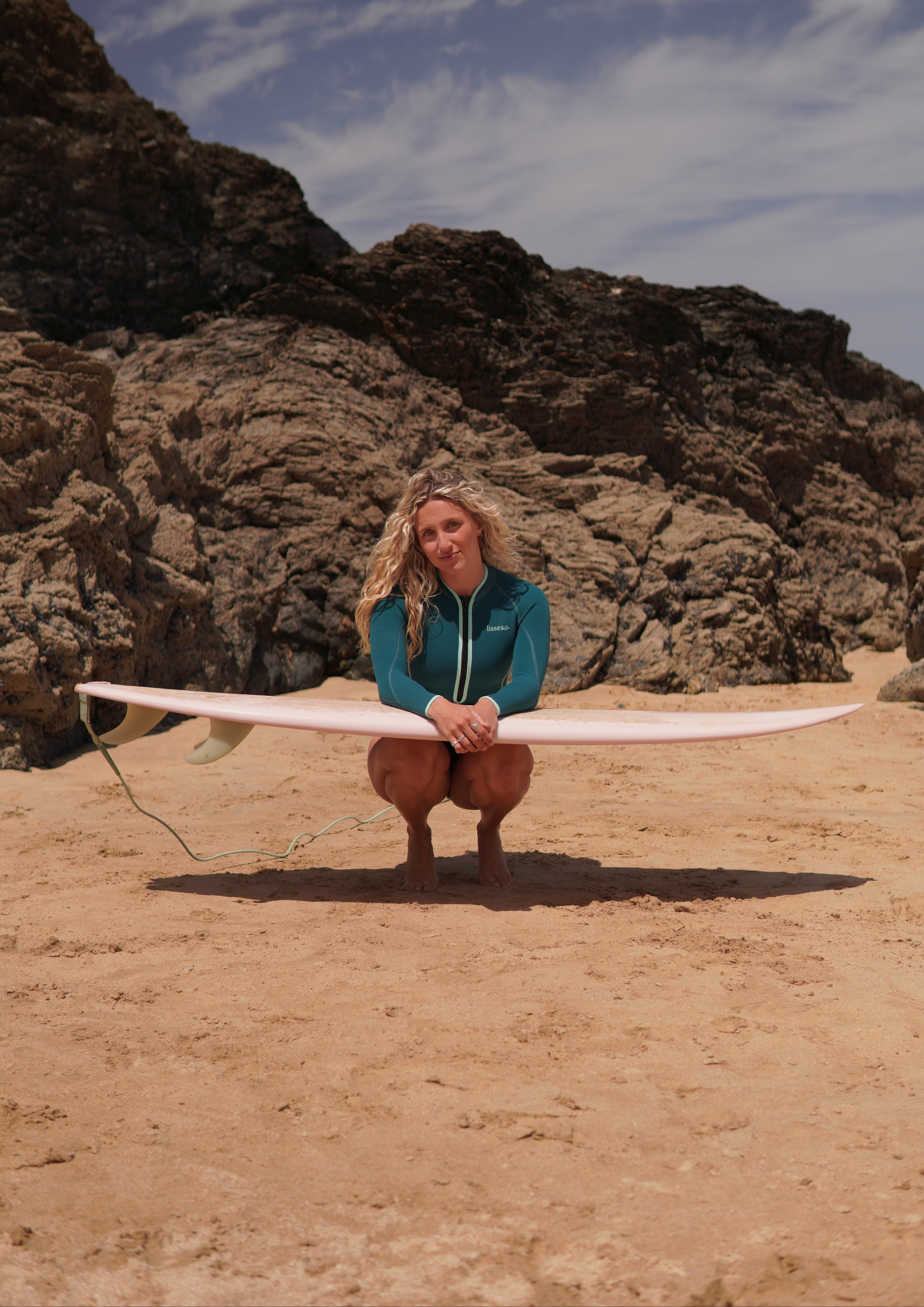 Woman in teal springsuit holding pink surfboard on sandy beach with rocky cliffs