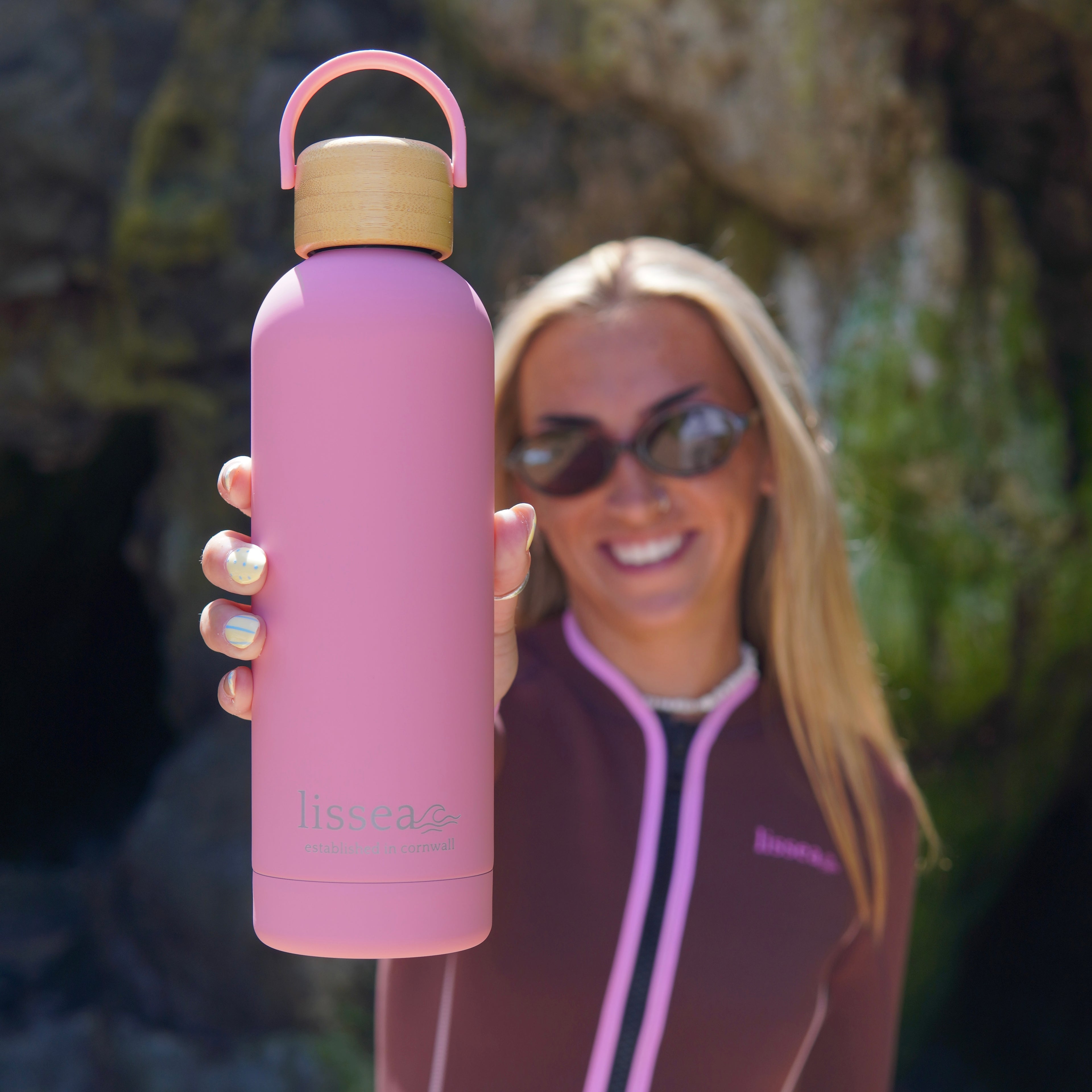 Pink Lissea water bottle with bamboo lid, held by woman in springsuit near rocky coast