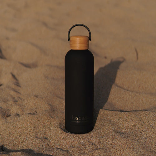 Black reusable water bottle with bamboo lid on sandy beach, perfect for ocean adventures and beachwear essentials.