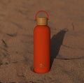 Orange reusable water bottle with bamboo lid on sandy beach, perfect for ocean adventures and beachwear essentials.