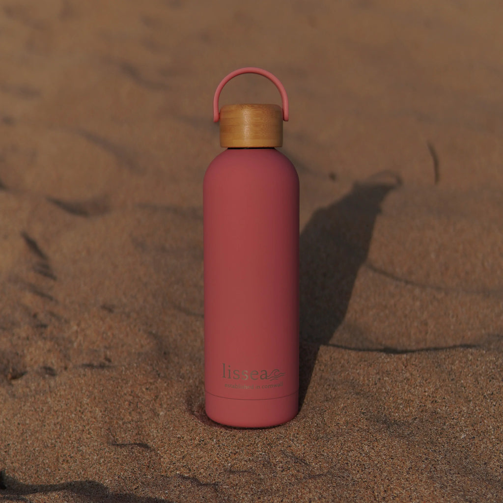 Pink Lissea water bottle with bamboo lid on sandy beach, perfect for ocean adventures and beachwear essentials.
