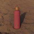 Pink Lissea water bottle with bamboo lid on sandy beach, perfect for ocean adventures and beachwear essentials.