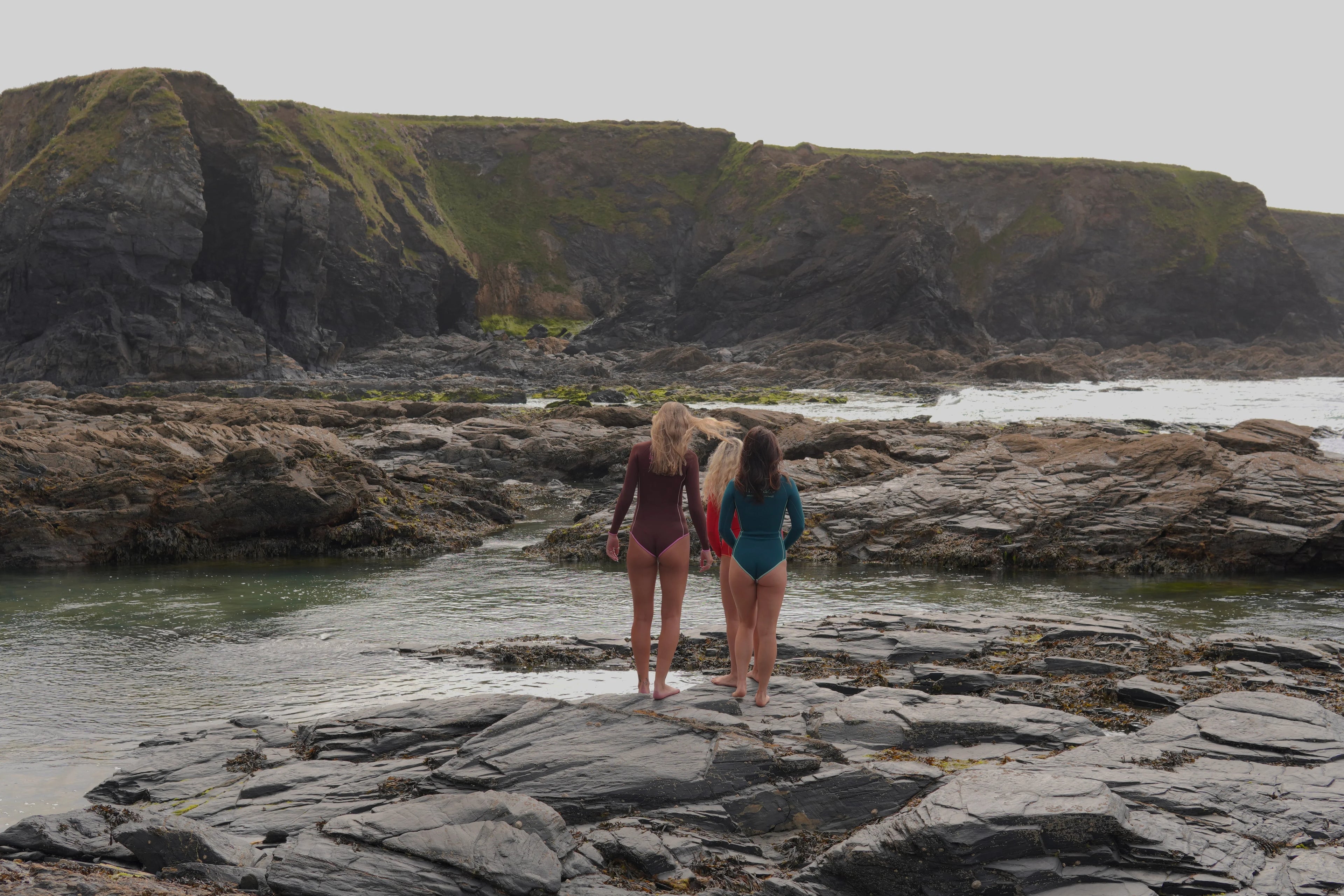 Women in long sleeve springsuits standing on rocky shore, ocean view, coastal adventure wear