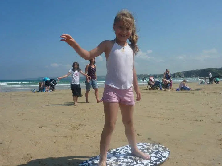 Girl standing on surfboard at beach wearing white top and pink shorts, enjoying ocean adventure