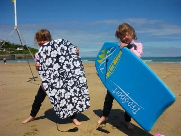Two kids on beach holding floral and blue bodyboards, ready for ocean fun with surf gear.