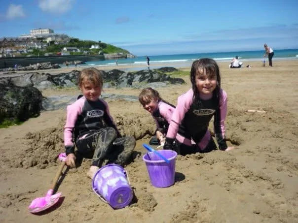 Kids in pink and black springsuits playing with buckets and shovels on sandy beach near ocean